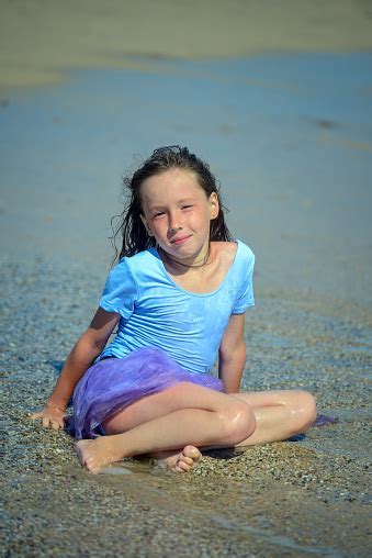 A Cute Little Girl In A Purple Swimsuit Sits On The Wet San