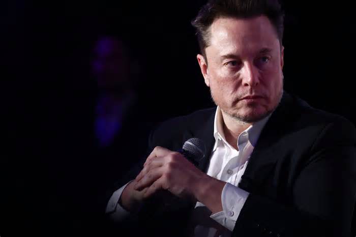 Elon Musk is looking to the right pensively with a microphone in his hand in front of a black background.