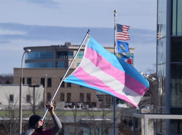 A protester holds a transgender pride flag in downtown Sioux Falls with the American, South Dakota and Sioux Falls flags flying in the near distance. They were with more than three dozen protestors who stood at Van Eps Park as part of a 