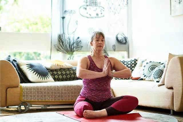 Person in a pink tank top and leggings sitting cross-legged in their living room, meditating to help lower blood pressure.