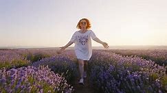 Redhaired woman runs forward joyfully through lavender field smiling in white dress touching blooming lavender flowers with her hands on walk in summer sunset. Lifestyle. Relax freedom. Aromatherapy
