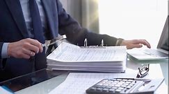 Businessman using a calculator and magnifying glass while analyzing financial documents at desk in fair modern office. Audit and taxes in business