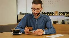 Стоковое видео категории «Бесплатно» — Хипстер в очках использует кредитную карту для онлайн-покупок со смартфоном, сидя в кафе